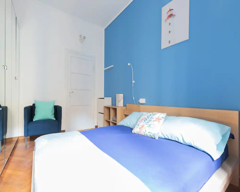 Camera da letto moderna e luminosa con letto matrimoniale con biancheria blu, testiera in legno, poltrona accogliente e armadio con ante specchiate — perfetta per un annuncio che mette in risalto uno spazio notte confortevole e ben illuminato.