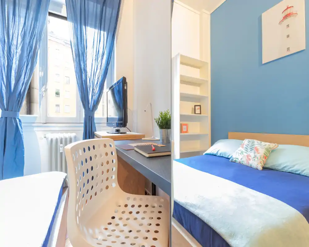 Camera da letto luminosa e compatta con letto ben rifatto, parete blu e angolo studio vicino alla finestra con scrivania e sedia — ideale per studenti o single.