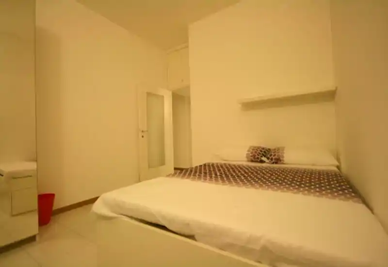 Minimalist bedroom with a double bed pushed against the wall, patterned runner and cushions, a small shelf above the headboard and a mirrored wardrobe visible at left. Clean, simple decor with warm lighting — ideal for short stays.