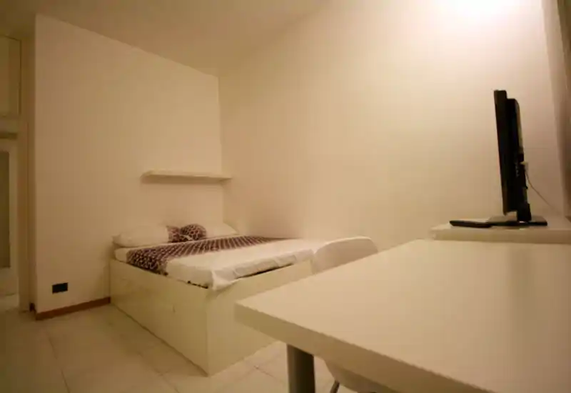 Minimalist double bedroom with a low white bed, patterned bedding and a small desk with TV; clean, simply furnished and well lit.