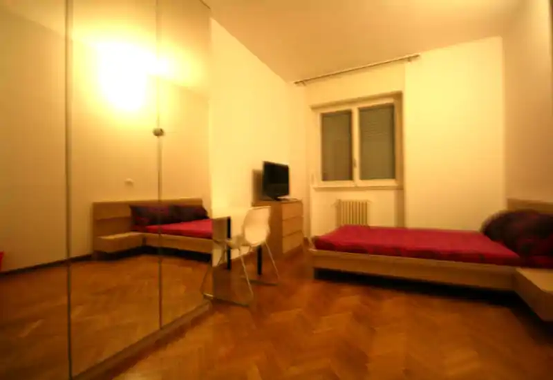 Camera da letto semplice con pavimento in legno a spina di pesce, un letto basso con biancheria rossa, un piccolo comò con TV e un armadio a specchio che riflette la stanza. L'illuminazione è calda e l'arredamento è minimal e funzionale.