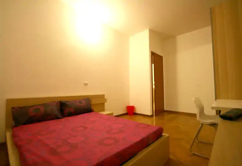 Camera da letto luminosa e ordinata con letto matrimoniale con biancheria rossa a fantasia, testiera semplice, scrivania e sedia. Illuminazione calda mette in risalto il parquet e l’arredo moderno e minimal.