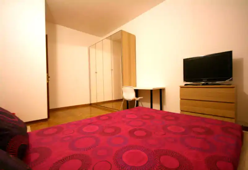 Ampia camera da letto con grande letto e copriletto rosso a motivo, armadio con ante a specchio, piccola scrivania e TV su comò in legno — ambiente caldo e ben sistemato.