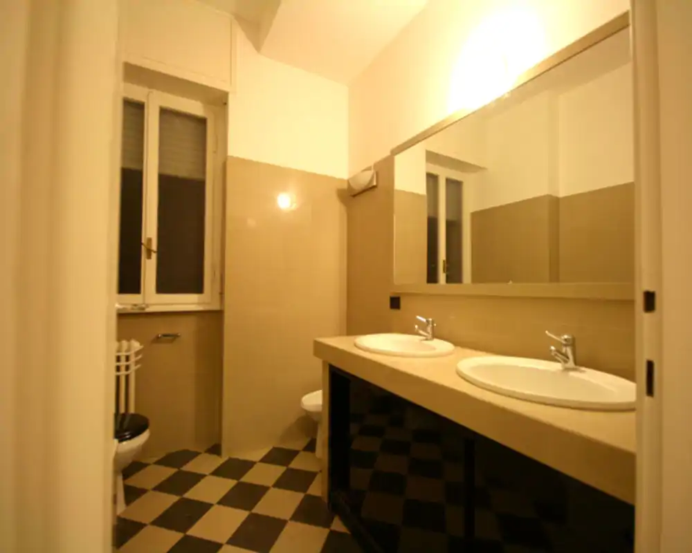 Bagno luminoso con doppi lavabi, specchio grande, pareti piastrellate e pavimento a scacchi — ambiente funzionale e pulito con luce naturale dalla finestra.