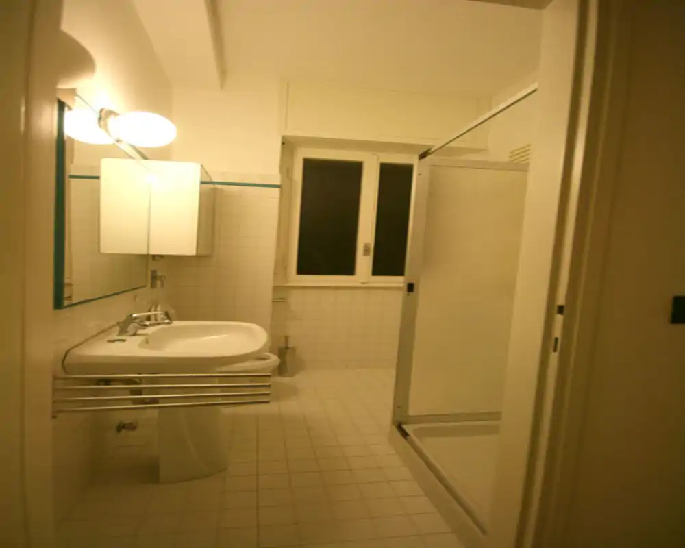 Bagno compatto con lavabo a colonna, mobile specchiato, lampada a parete e cabina doccia angolare; piastrelle bianche e una finestra alta per ventilazione naturale.