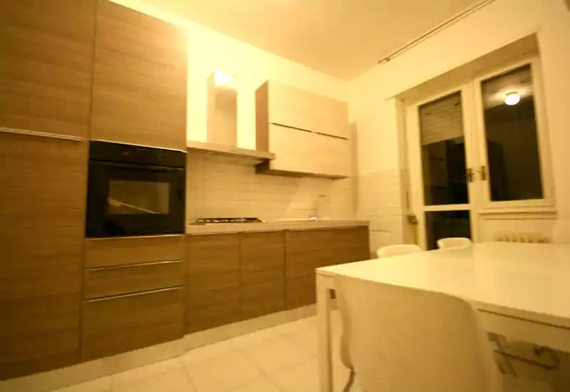 Cucina moderna ben illuminata con mobili effetto legno, forno integrato, piano cottura e tavolo da pranzo in primo piano — uno spazio funzionale e adatto alla vita quotidiana.