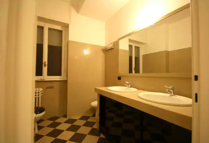 Bagno luminoso con doppi lavabi, specchio grande, pareti piastrellate e pavimento a scacchi — ambiente funzionale e pulito con luce naturale dalla finestra.