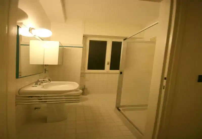 Compact bathroom with a pedestal sink, mirrored cabinet, wall lamp and a corner shower cubicle; neutral white tiles and a tall window provide natural ventilation.