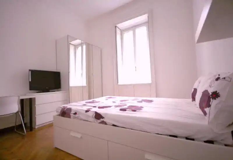 Chambre lumineuse et minimaliste avec lit double habillé d'une parure florale, armoire miroir, petit bureau et télévision — propre, moderne et bien éclairée par une grande fenêtre.