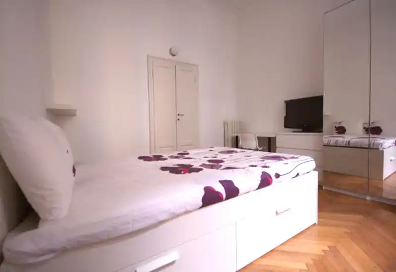 Chambre lumineuse et minimaliste avec lit blanc à couette florale, armoire miroir et parquet. Style épuré et bonne luminosité, adaptée aux annonces locatives.