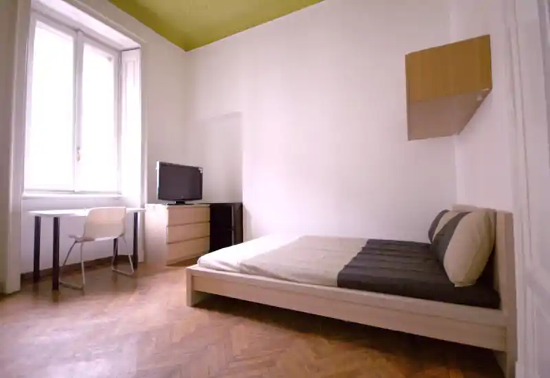 Bright, minimal bedroom with a low wooden bed, neutral linens and a large window providing natural light. The room includes a small desk and dresser, perfect for a single occupant or student.
