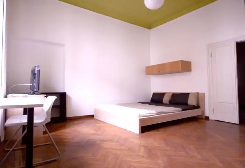 Minimal, bright bedroom with a low wooden double bed, neutral bedding, a wall-mounted cabinet and a desk by the window; wooden parquet floor and high ceiling create an airy feel.