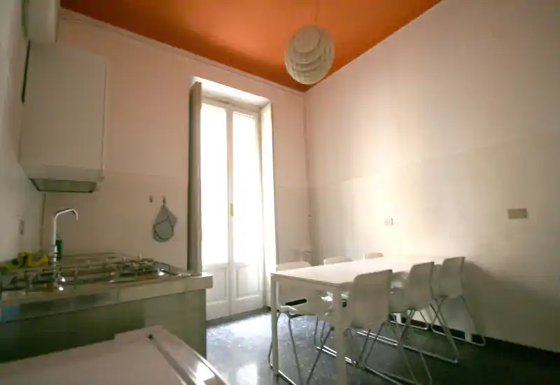 Bright kitchen with stainless-steel counter and a simple white dining table with chairs beside a tall window/door; high ceiling with warm orange paint adds character.