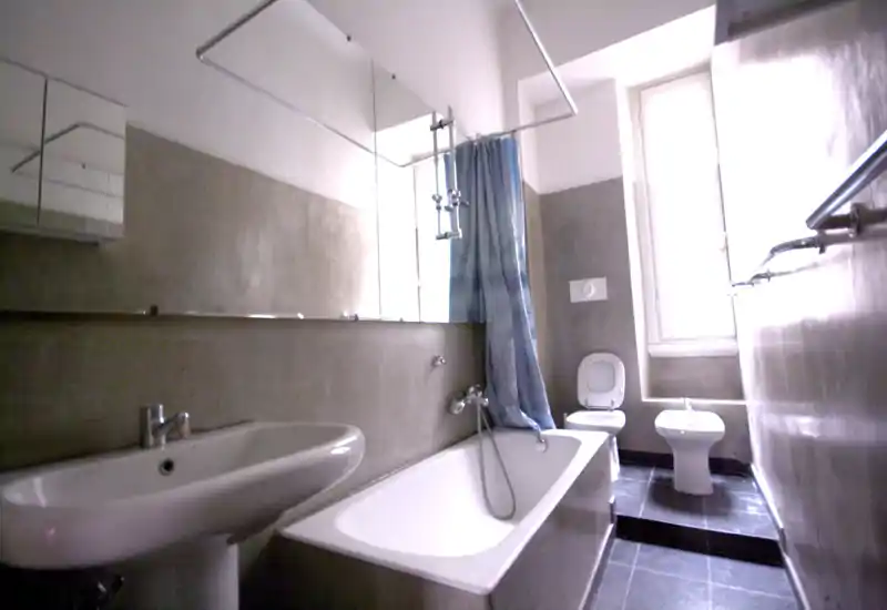 Salle de bains lumineuse avec baignoire, lavabo, toilettes et bidet ; grand miroir et fenêtre apportent de la lumière naturelle dans un agencement compact et fonctionnel.