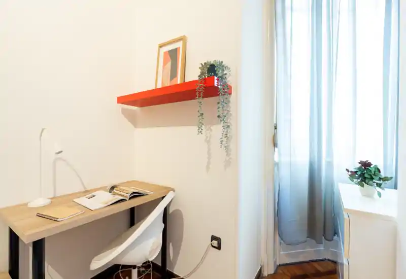 Coin bureau minimaliste et lumineux avec petit bureau, lampe de travail, chaise et étagère décorative. La lumière naturelle filtre à travers des rideaux légers, créant un coin travail propre et accueillant.