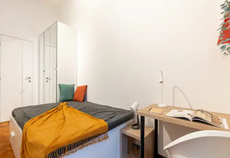 Chambre compacte et lumineuse avec un lit simple bien fait, jeté et coussins colorés, un petit bureau et une chaise — idéale pour un étudiant ou une personne seule.