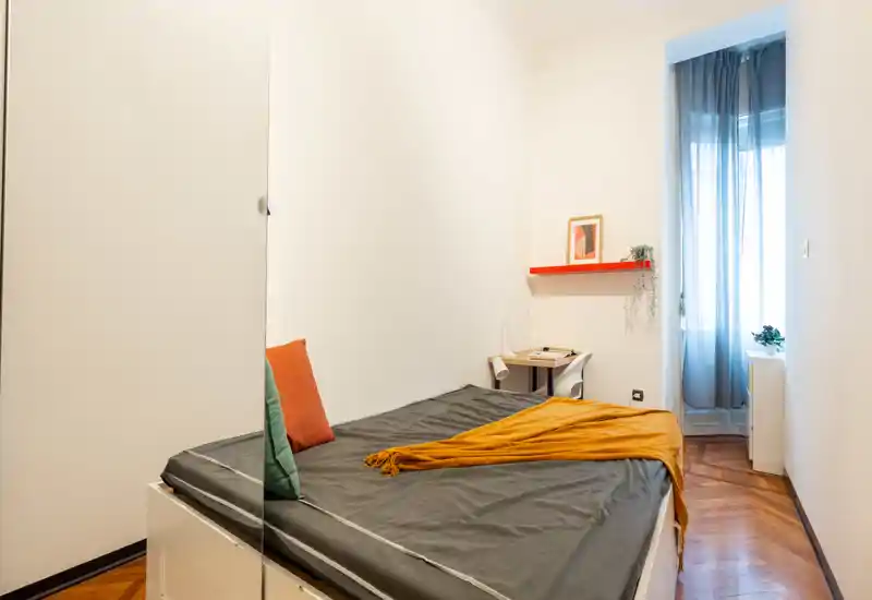 Chambre compacte et lumineuse avec un lit double habillé de linge sombre, coussins colorés et un plaid moutarde. La lumière naturelle entre par une grande fenêtre à voilages; un petit bureau et une étagère ajoutent du caractère fonctionnel.