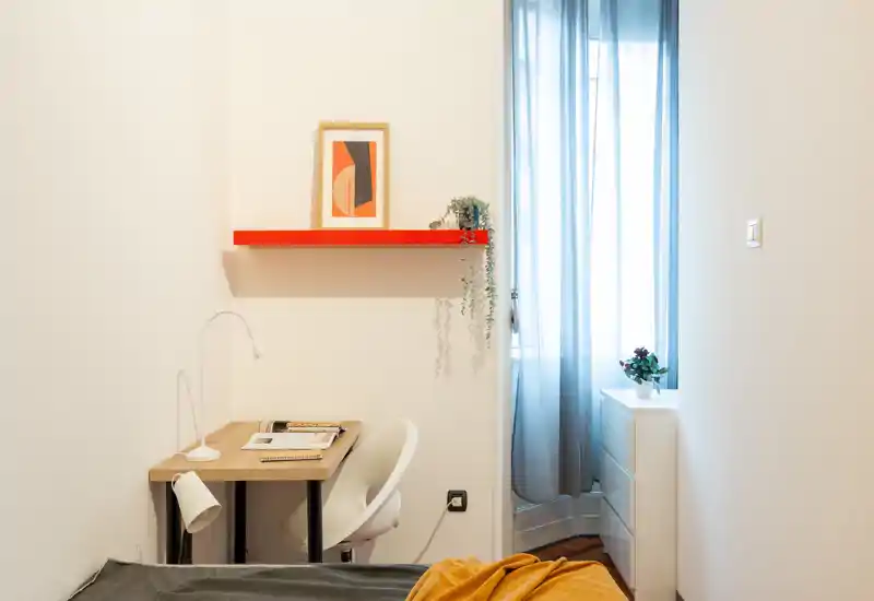 Coin chambre compact et lumineux avec lit au premier plan, petit bureau et chaise, et une haute fenêtre aux rideaux légers laissant passer une douce lumière naturelle.