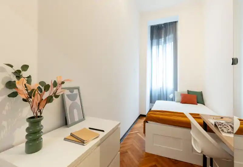 Dormitorio compacto y con estilo con cama individual, ropa de cama cálida, un pequeño escritorio y luz natural de una ventana alta — ideal para un alquiler acogedor.