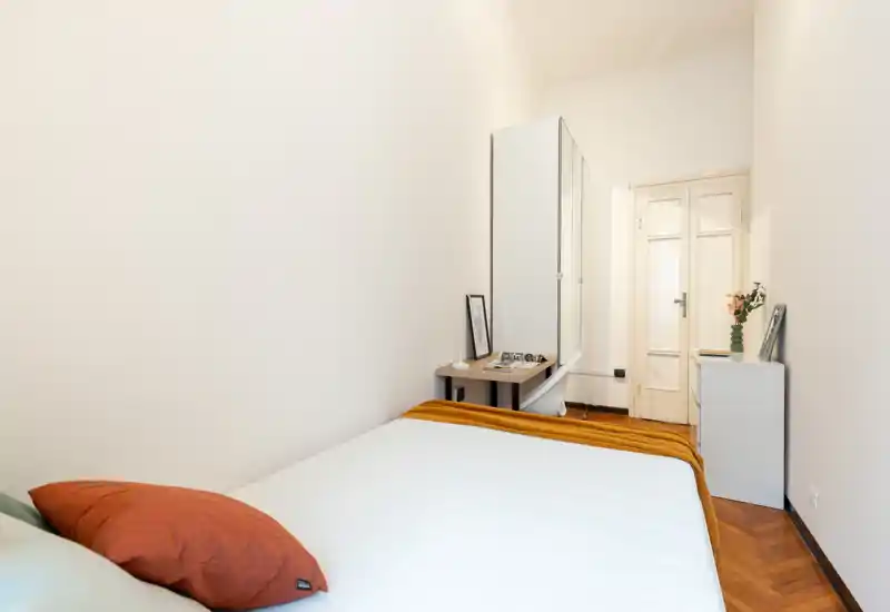 Dormitorio luminoso y minimalista con cama bien hecha, manta cálida y cojines; espejo alto y pequeño escritorio junto a la puerta para vestirse y guardar.
