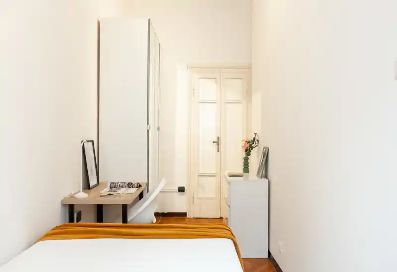 Dormitorio luminoso y minimalista con cama bien hecha, pequeño escritorio y armario. Una manta cálida y una decoración sencilla hacen el espacio acogedor y funcional, ideal para una persona.