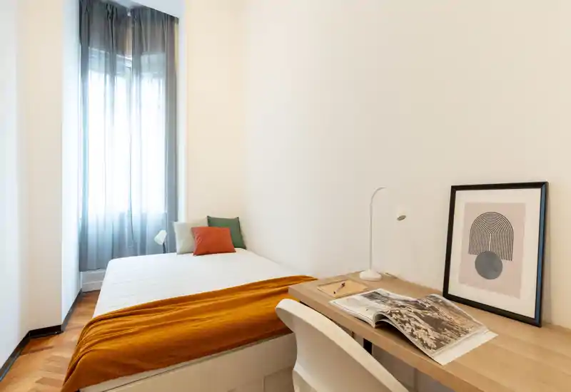 Habitación individual minimalista y luminosa con cama bien arreglada, manta naranja cálida y cojines decorativos, además de un escritorio compacto con lámpara y marco: ideal para estudiantes o alquileres cortos.