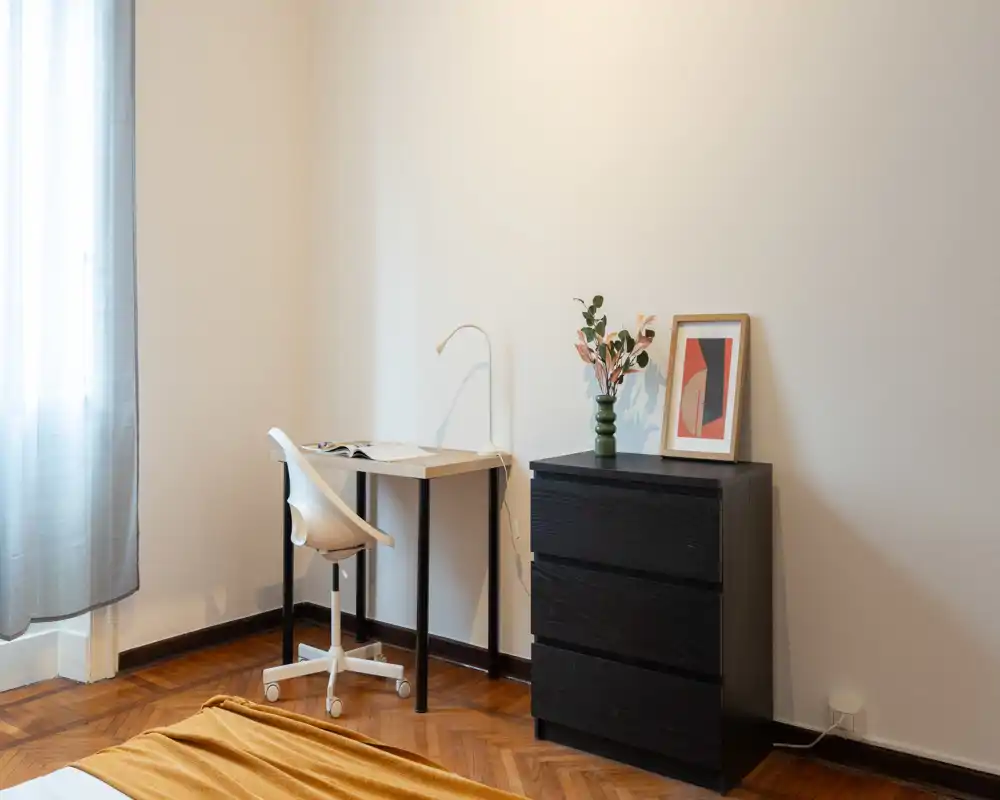 Angolo camera minimalista e luminoso con una scrivania piccola e un comò nero a tre cassetti; luce naturale morbida e pavimento in legno caldo.