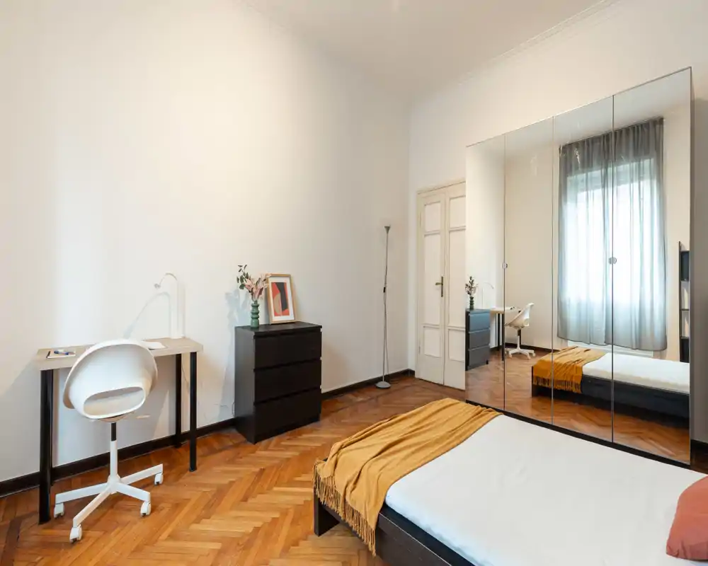 Camera da letto luminosa e minimalista con letto basso, plaid color senape e un grande armadio con ante a specchio che amplia lo spazio. Una piccola scrivania e un comò vicino alla finestra creano un angolo pratico per lavorare.