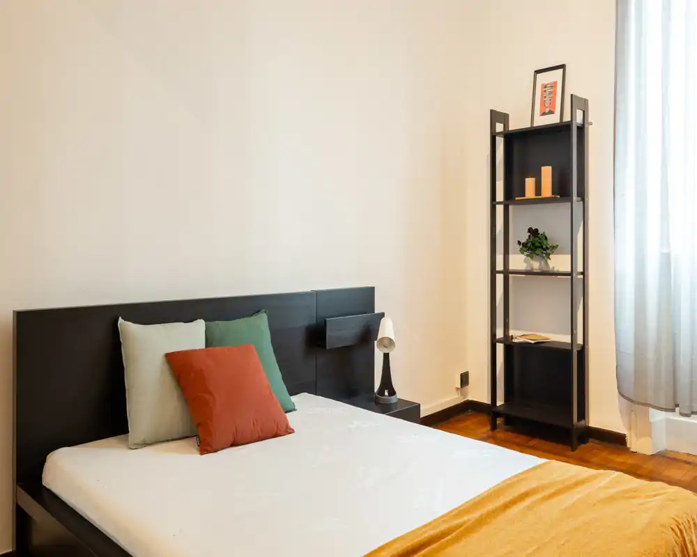 Camera da letto luminosa e moderna con testiera in legno scuro, materasso ordinato, cuscini colorati e una sottile libreria vicino alla finestra.