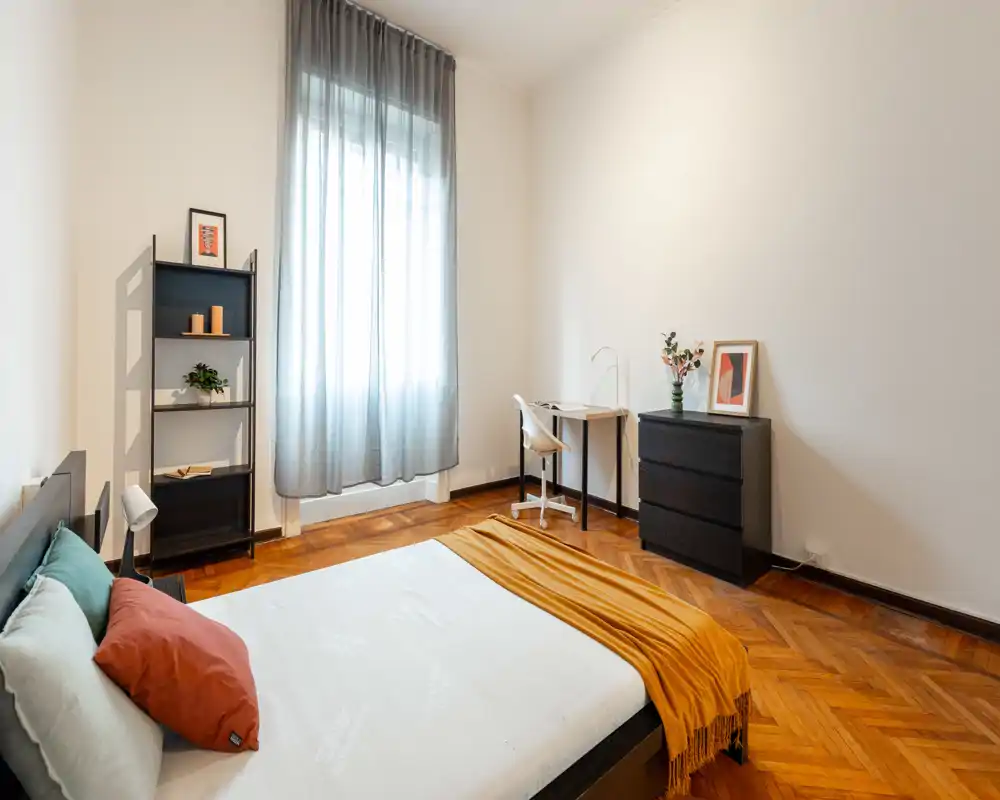 Camera da letto luminosa e ben arredata con letto matrimoniale, pavimento in legno caldo e finestra che diffonde luce naturale; scrivania e comò offrono spazio di lavoro e contenimento.