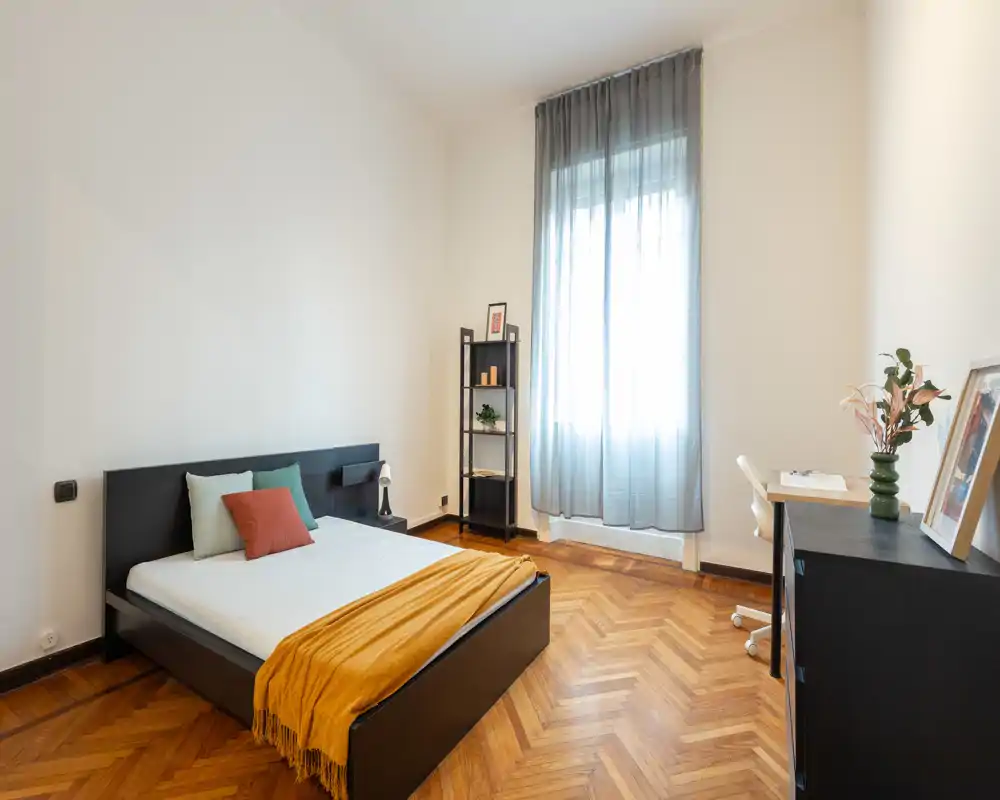 Camera da letto luminosa e minimal con letto basso nero, pavimento in parquet a spina di pesce e scrivania vicino alla finestra. Tessili e cuscini aggiungono comfort mentre la luce naturale illumina l’ambiente.