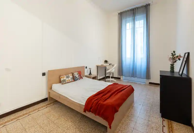 Camera da letto semplice e luminosa con letto in legno, cuscini decorativi e un plaid rosso. Una scrivania e una sedia sono posizionate vicino a una finestra alta con tende leggere, completa di un piccolo comò nero e un vaso.