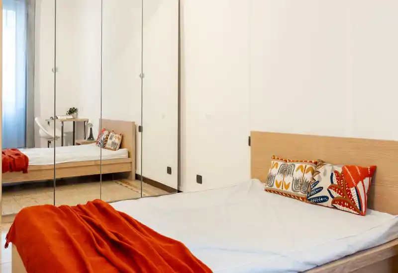 Camera da letto moderna e luminosa con letto in legno, cuscini decorativi e un caldo plaid arancione; la guardaroba con ante a specchio amplia la stanza e si intravede una scrivania nel riflesso.