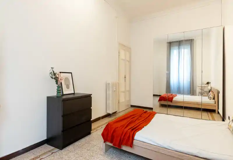 Camera da letto luminosa e minimale con un letto singolo in legno, plaid rosso e un armadio con specchi che riflette la finestra. Linee pulite, arredo semplice e luce naturale la rendono accogliente e facile da allestire.