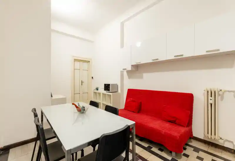 Zona de estar/comedor bien iluminada con mesa moderna de cristal, cuatro sillas y un sofá rojo llamativo. Paredes blancas y armarios altos aportan un espacio luminoso y ordenado, perfecto para alquileres temporales.
