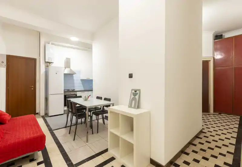 Zona de estar compacta tipo open-plan con mesa de comedor, sofá rojo y cocina pequeña al fondo — bien iluminada y amueblada de forma sencilla para alquileres urbanos.