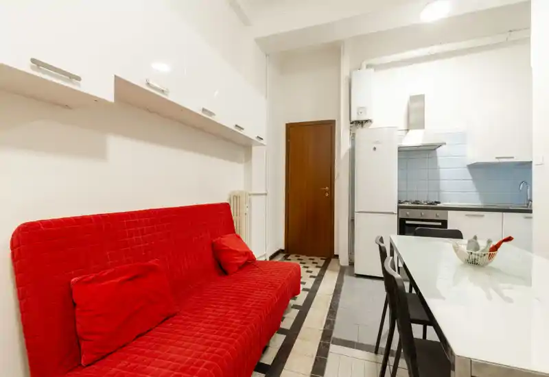Zona de estar compacta y luminosa con un sofá rojo llamativo, muebles altos y una pequeña cocina abierta con mesa — ideal para un estudio acogedor.