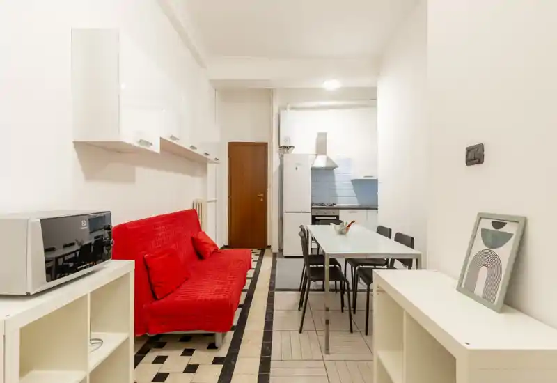 Zona de estar de planta abierta con sofá rojo, mesa de comedor y pequeña cocina al fondo. Espacio luminoso y limpio con muebles blancos modernos y detalles decorativos sencillos.