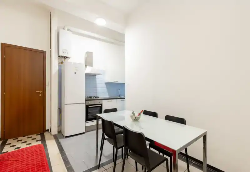 Cocina compacta con horno a gas, frigorífico y mesa de comedor de cristal con cuatro sillas; paredes blancas y salpicadero de azulejos azules que ofrecen un espacio limpio y funcional.