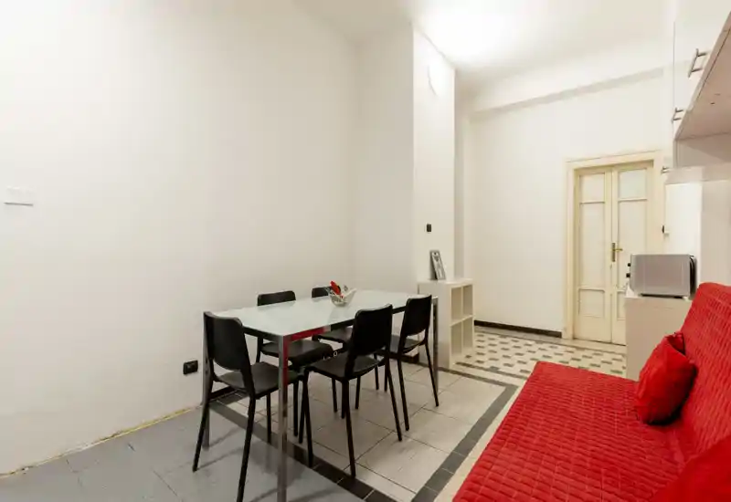 Zona de comedor luminosa y sencilla con mesa de cristal y seis sillas, además de un banco acolchado rojo; paredes neutras y suelo de baldosas para un espacio limpio y funcional.
