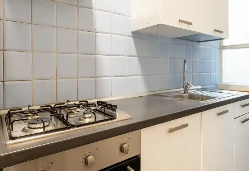Cocina compacta y funcional con placa de gas, fregadero de acero inoxidable y salpicadero de azulejos claros; luz natural y muebles blancos sencillos.