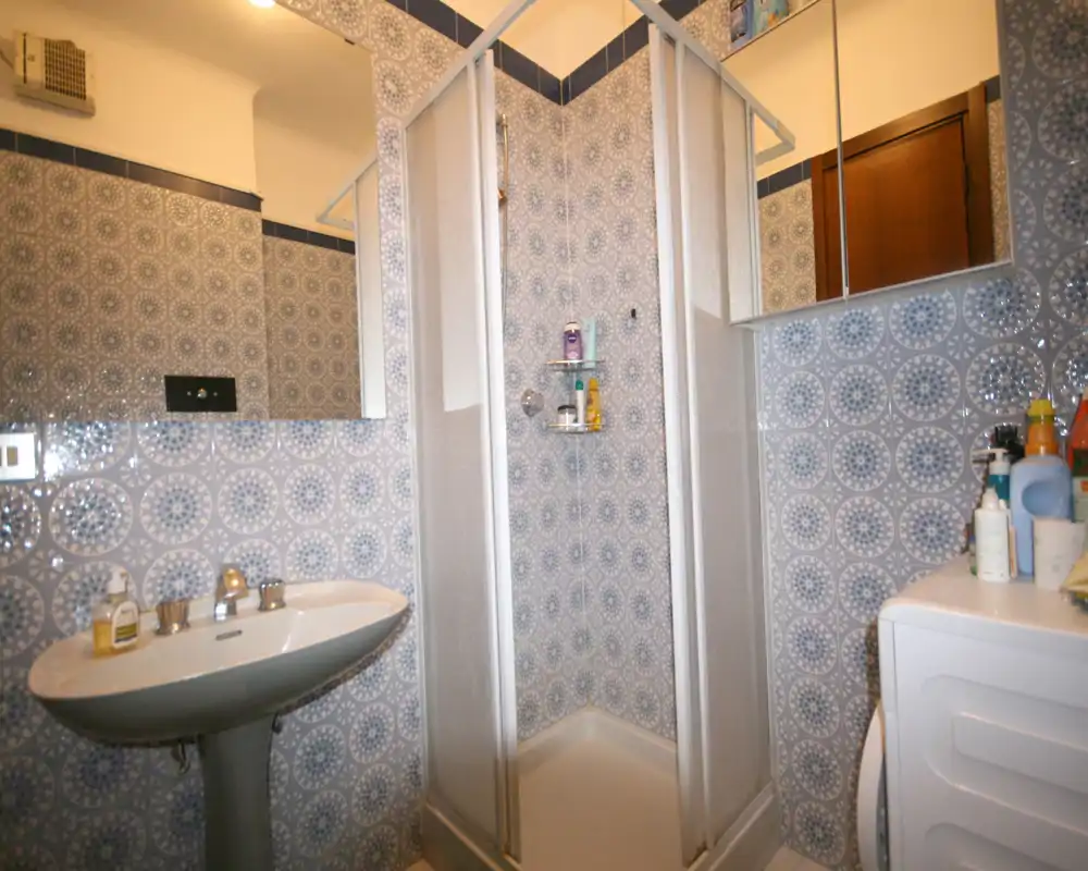 Baño compacto con cabina de ducha en esquina, lavabo de pie y lavadora. Las paredes están revestidas con azulejos azules decorativos y hay un espejo armario sobre el lavabo.