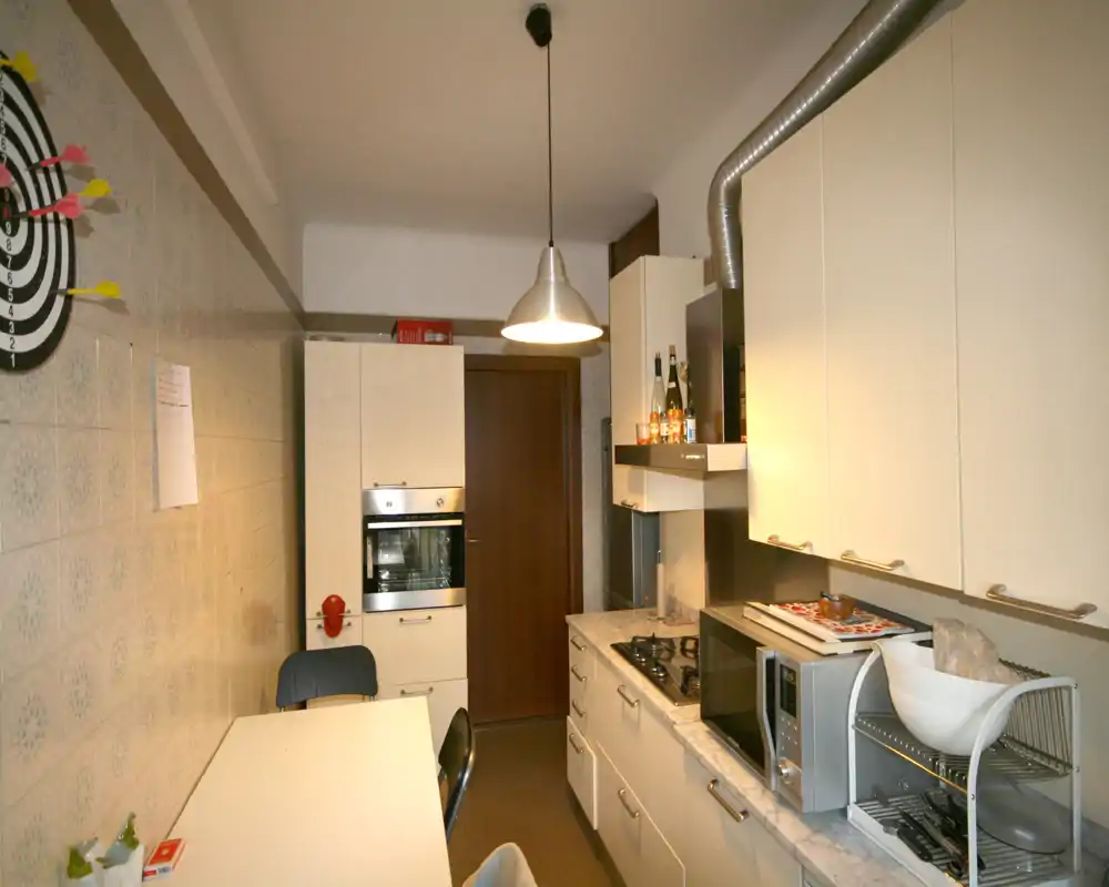 Cocina estrecha y luminosa con armarios blancos, encimera efecto mármol, horno empotrado y vitro a gas, y una superficie práctica para comer — espacio funcional y ordenado.