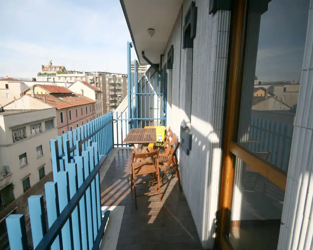 Balcón soleado y estrecho con pequeña mesa y sillas de madera, barandilla azul y vistas urbanas — ideal para el café matutino.