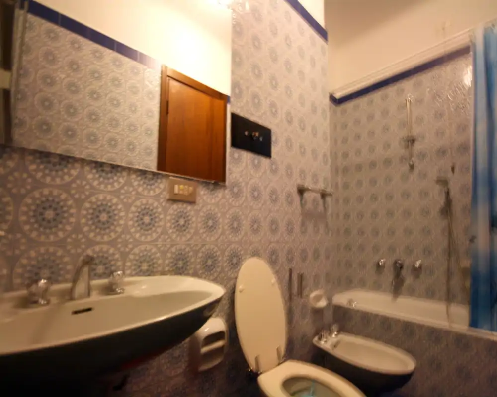 Baño con azulejos azules estampados, lavabo, inodoro, bidé y bañera con ducha; imagen ligeramente desenfocada y luz cálida.