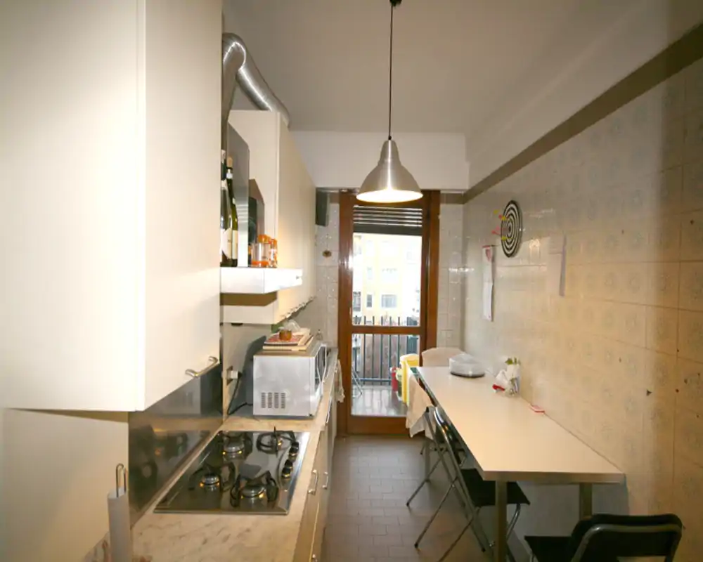 Cocina en galería con placa de gas, pequeños electrodomésticos, mesa comedor y puerta a un pequeño balcón. Lámpara colgante y azulejos en la pared dan un aspecto práctico y acogedor para el uso diario.