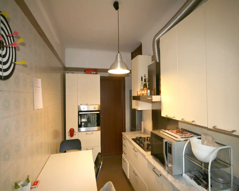 Cocina en galería bien equipada con muebles blancos, horno empotrado, placa de gas y electrodomésticos sobre la encimera; lámpara colgante luminosa y superficies ordenadas, ideal para preparar comidas.