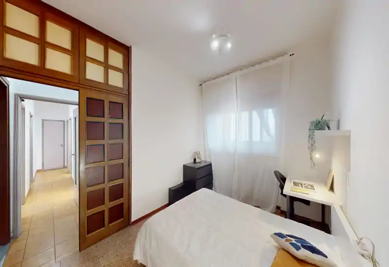 Compact, tidy bedroom with a double bed, natural light from a curtained window, a small desk and chest of drawers—ideal for a single occupant or couple.