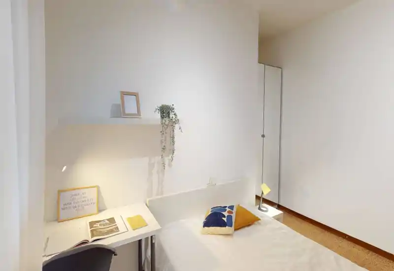 Bright, minimal bedroom with a single bed, decorative cushions, a small desk and warm task lighting — neat and welcoming for a solo guest.