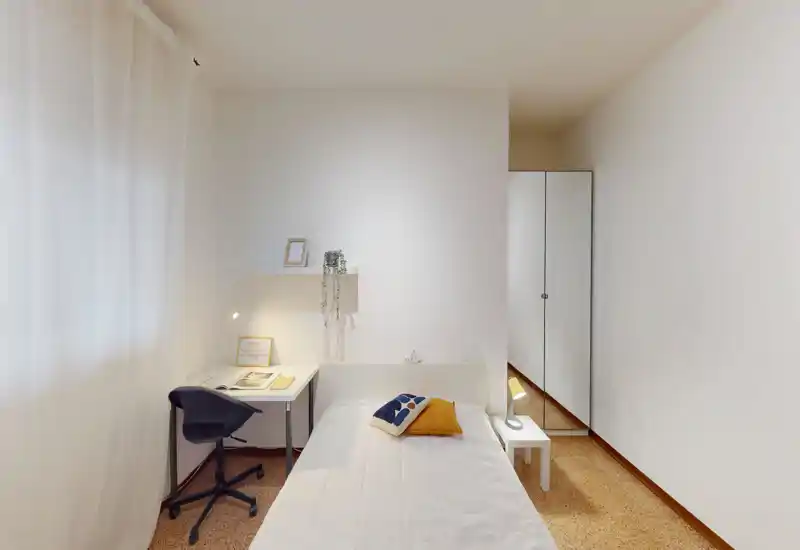 Bright, minimalist bedroom with a single bed, bedside table and a small desk by the window. Clean lines, soft natural light and simple decorative cushions create a calm, inviting atmosphere.